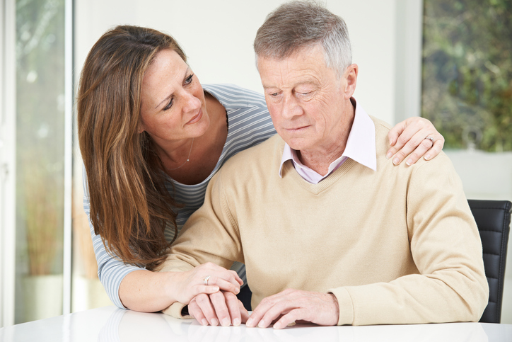 A concerned looking woman with long brown hair crouches down to put her arm around a sitting elderly man who appears sad